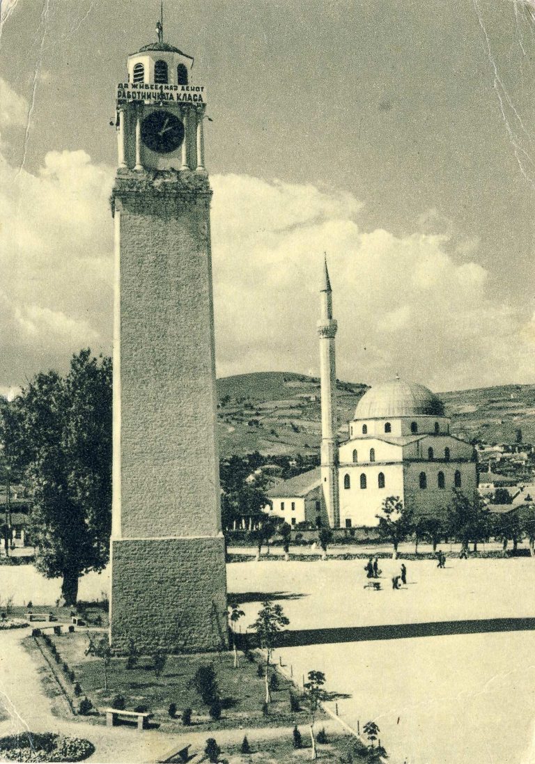 Изглед на Саат ку­лата во новоформираниот голем плоштад во месец мај 1954 година. Високо на огра­дата стои платно со по­рака: Да живее 1-ви Мај денот на работничката класа. Во заднината се гледа Исак џамијата и ридот на север­ниот дел наречен Смилевски Баир. Околу Саат кулата е новоформираниот парк со млади дрвја.